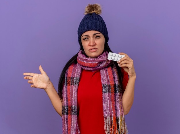 Schwaches junges kaukasisches krankes Mädchen, das Wintermütze und Schal trägt, die Packung der Tabletten betrachten Kamera betrachten, die zur Seite mit Hand lokalisiert auf lila Hintergrund mit Kopienraum zeigt