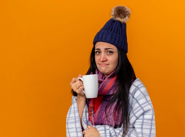 Schwaches junges kaukasisches krankes Mädchen, das Wintermütze und Schal trägt, eingewickelt in Plaid, das Tasse Tee hält und Kamera lokalisiert auf orange Hintergrund mit Kopienraum betrachtet