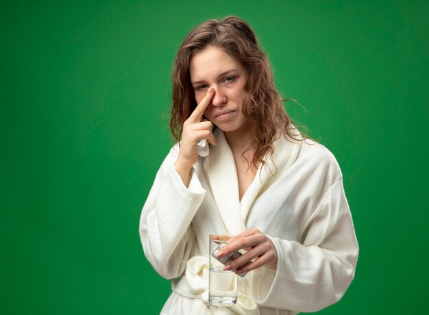 Schwaches junges krankes Mädchen, das weißes Gewand trägt, das Glas Wasser mit Pillen hält, die Finger auf Nase lokalisiert auf Grün mit Kopienraum setzen