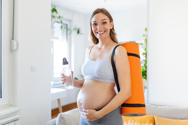 Schwangere Frau, die eine Yogamatte und eine wiederverwendbare Wasserflasche hält, bereitet sich darauf vor, zu Hause zu trainieren Wohlbefinden Während der Schwangerschaft fit und gesund bleiben