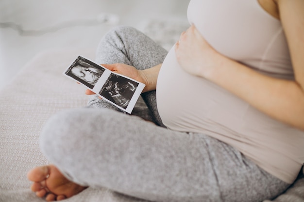 Schwangere Frau mit Ultraschallfoto auf dem Bett sitzend