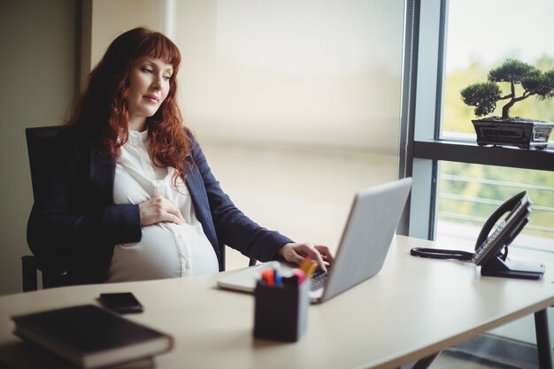 Schwangere Geschäftsfrau, die ihren Bauch hält, während Laptop verwendet