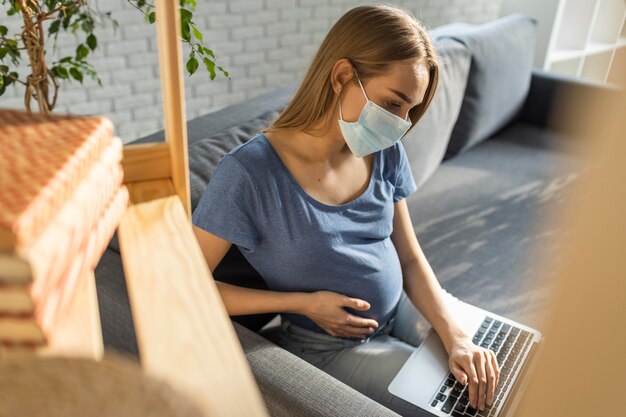 Schwangere Geschäftsfrau mit medizinischer Maske, die auf Couch sitzt und am Laptop arbeitet