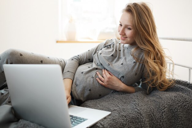 Schwangerschaftszeit mit Freude verbringen. Schöne blonde junge schwangere Frau in der bequemen Hauptkleidung, die auf Bett liegt, auf der Suche nach einem guten Film ist, um auf Laptop zu sehen, sich zu entspannen, Hand auf ihrem Bauch zu halten.