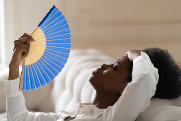Schwarze geschäftsfrau, die an hitzschlag leidet, sitzt zu hause im wohnzimmer mit einem winkenden ventilator
