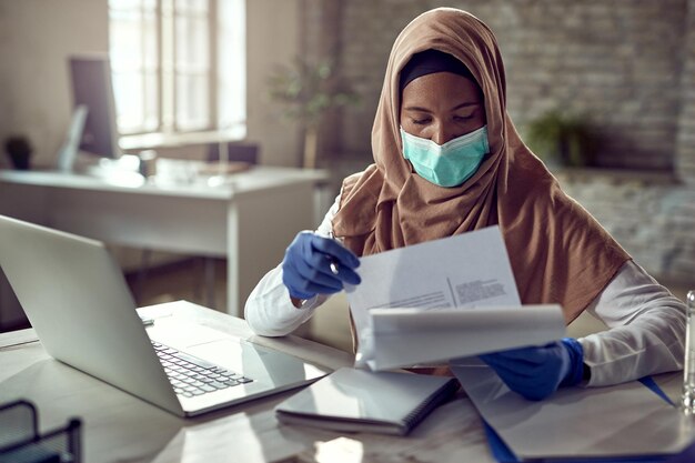 Schwarze islamische Geschäftsfrau mit schützender Gesichtsmaske, während sie während einer Viruspandemie im Büro Papierkram durchgeht