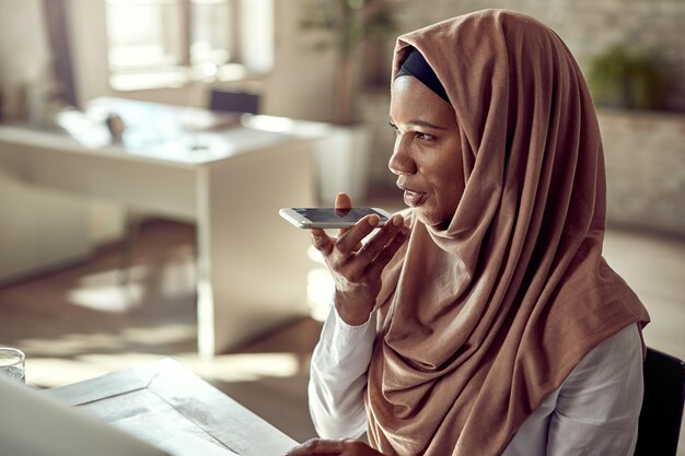 Schwarze muslimische Unternehmerin nimmt während der Arbeit in ihrem Büro Sprachnachrichten auf dem Handy auf