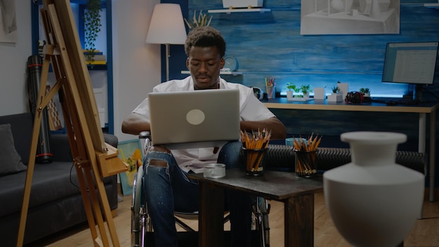 Schwarze person im rollstuhl mit laptop-computer für kunstinspiration, die im kreativitätsraum sucht. behinderter afroamerikanischer künstler, der professionelle meisterwerke im modernen studio entwirft