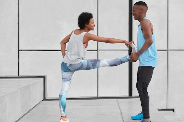 Schwarze positive männliche ausbilder helfen weiblichen auszubildenden, übungen für flexibilität zu machen, stehen an treppen an der weißen wand, haben glückliche gesichtsausdrücke und sind in sportbekleidung gekleidet. menschen-, sport- und trainingskonzept