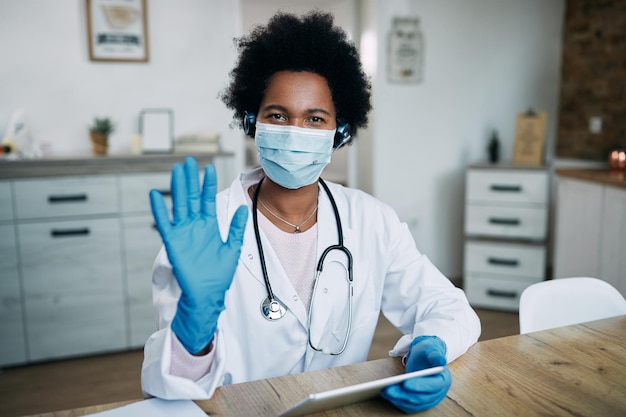 Schwarze Ärztin mit schützender Gesichtsmaske und Handschuhen mit Touchpad und winken zur Kamera