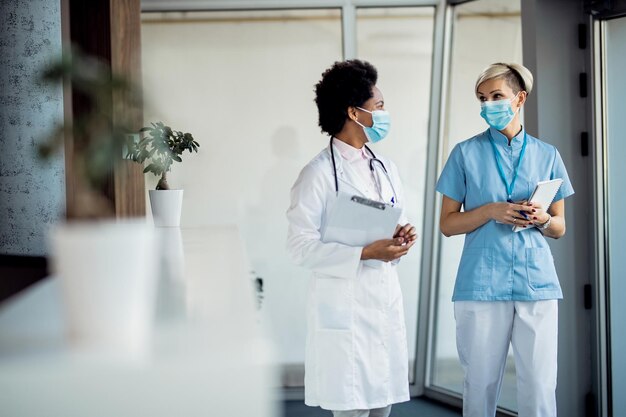 Schwarze Ärztin und Krankenschwester kommunizieren, während sie Gesichtsmasken tragen und durch den Krankenhausflur gehen