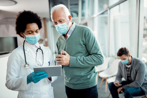 Schwarze Ärztin und Seniorin mit Touchpad beim Tragen von Gesichtsmasken in der medizinischen Klinik