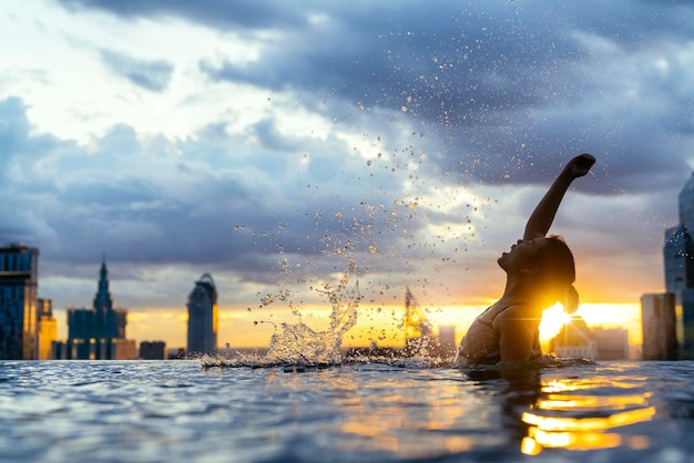 Schwarze Silhouette einer asiatischen Frau Spritzwasser in den Sommerferien, die sich im Infinity-Pool mit Blick auf den Sonnenuntergang auf dem blauen Meer und der städtischen Innenstadt mit Hochhaus entspannen Gesunder Glückslebensstil