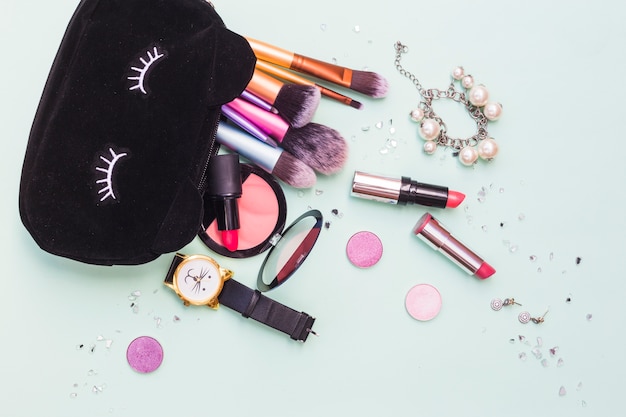 Schwarze Tasche mit Make-up Pinsel; Armband; Armbanduhr- und Kosmetikprodukt auf Pastellhintergrund