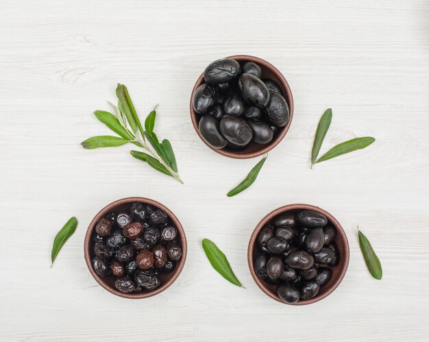 Schwarze und braune Oliven in Tonschalen mit Olivenbaumzweig und Draufsicht auf weißem Holz