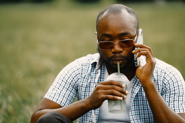 Schwarzer amerikanischer Mann, der auf einem Gras sitzt und am Telefon spricht. Mann mit weißem T-Shirt und blauem kariertem Hemd