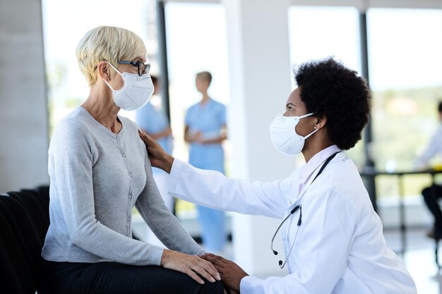 Schwarzer Arzt und reifer Patient mit Schutzmasken, während sie im Wartezimmer der Klinik sprechen