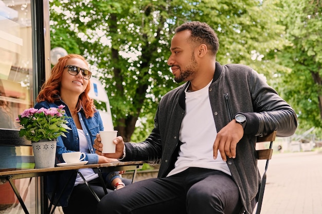 Schwarzer bärtiger Mann und rothaarige Frau trinken Kaffee in einem Café auf einer Straße.