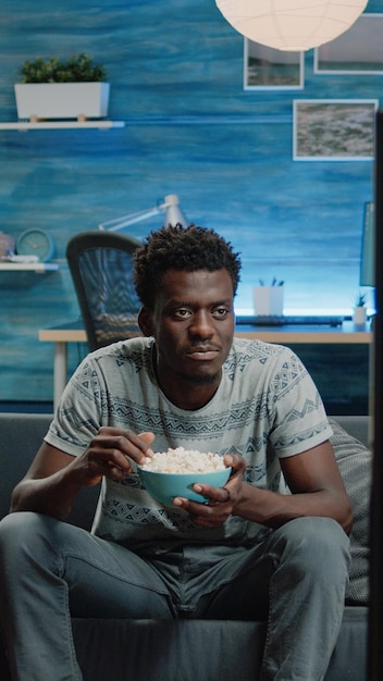 Schwarzer Erwachsener sieht Comedy-Film im Fernsehen, während er Popcorn aus der Schüssel isst. Afroamerikaner, der mit Snack lacht und fernsieht. Mann afrikanischer Ethnizität, der Spaß hat