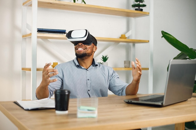 Schwarzer männlicher Unternehmer, der im Büro sitzt und eine Virtual-Reality-Brille verwendet