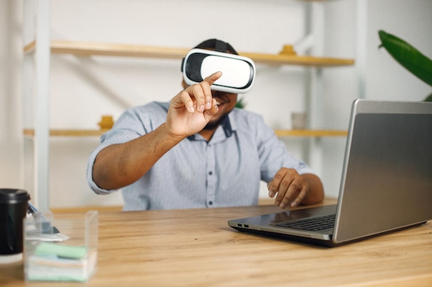 Schwarzer männlicher Unternehmer, der im Büro sitzt und eine Virtual-Reality-Brille verwendet