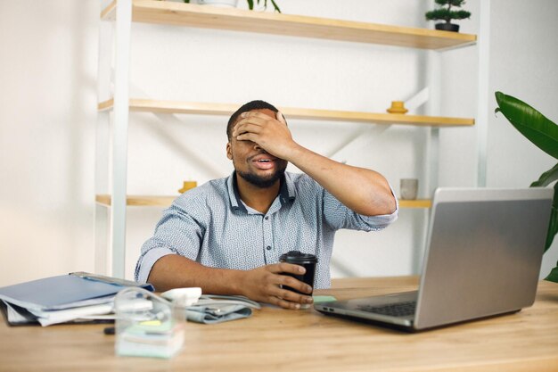 Schwarzer männlicher Unternehmer sitzt im Büro mit Laptop und trinkt Kaffee