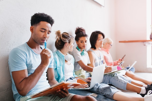 Kostenloses Foto schwarzer mann im blauen hemd, der versucht, mit schwierigem test herauszufinden. stilvolle studentin, die mit laptop auf dem boden sitzt und freunde ansieht und ihnen etwas erzählt.