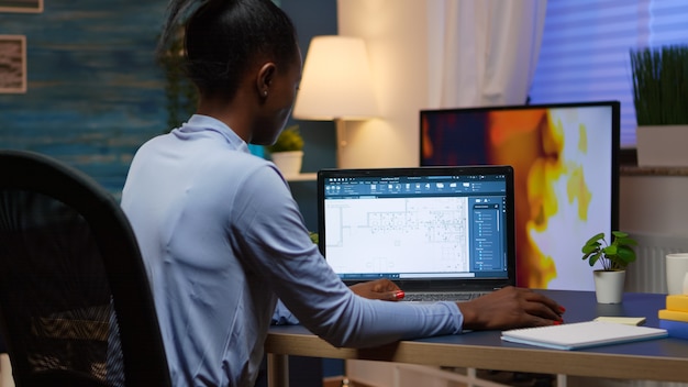 Schwarzer Mitarbeiter, der digitale Blaupausen überprüft, die das Projekt des Unternehmens analysieren, das auf dem Laptop sitzt, der spät in der Nacht am Schreibtisch im Wohnzimmerbüro sitzt. Vielbeschäftigter afrikanischer Freiberufler mit moderner Technologie