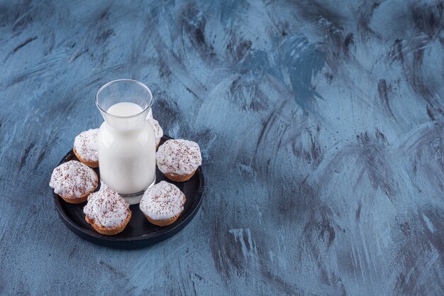 Schwarzer Teller mit süßen cremigen Cupcakes und einem Glas Milch auf Marmor.