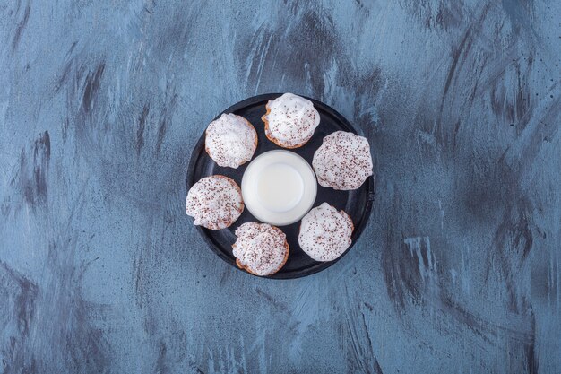 Schwarzer Teller mit süßen cremigen Cupcakes und einem Glas Milch auf Marmoroberfläche.