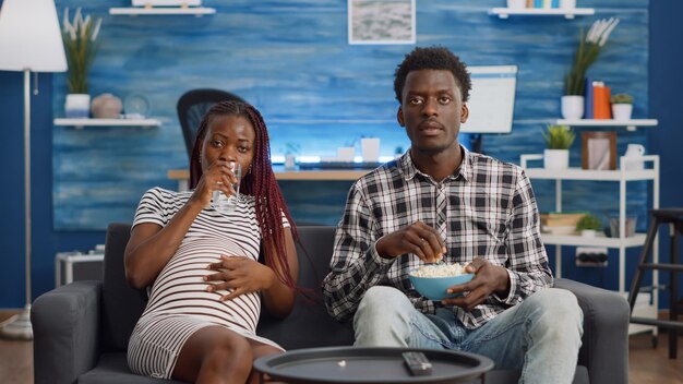 Schwarzes paar mit schwangerschaft entspannt sich zu hause vor dem fernseher im wohnzimmer. afrikanischer amerikanischer mann, der ein glas wasser für schwangere frau bringt, die popcorn auf der couch isst. verheiratete leute, die in die kamera schauen
