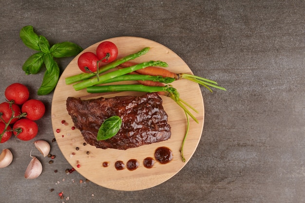 Schweinefleisch gebratene, gegrillte Spareribs von einem Sommergrill, serviert mit Gemüse, Spargel, Babykarotten, frischen Tomaten und Gewürzen. geräucherte Rippen auf Holzschneidebrett auf Steinoberfläche. Draufsicht.