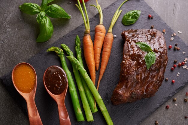 Schweinefleisch gebratene, gegrillte Spareribs von einem Sommergrill, serviert mit Gemüse, Spargel, Babykarotten, frischen Tomaten und Gewürzen. geräucherte Rippen auf schwarzer Steinoberfläche. Draufsicht,