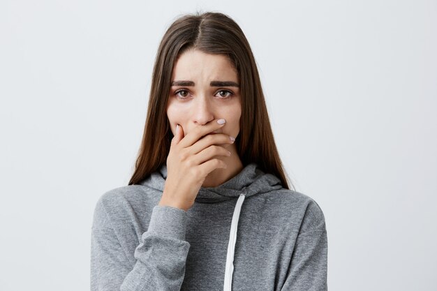 Schwierige Momente im Leben. Nahaufnahme des jungen schönen kaukasischen Mädchens mit langen Haaren im stilvollen grauen Kapuzenpulli, der weint und Hände nahe Mund hält, kann nicht glauben, dass ihr geliebter Opa gestorben ist.