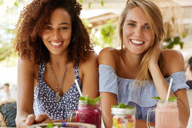 Schwule weibliche Paare haben positive Ausdrücke, sitzen in der Cafeteria nahe beieinander, lächeln freudig, genießen leckere Desserts in der Cafeteria im Freien. Multiethnische Lesben sprechen miteinander. Liebeskonzept