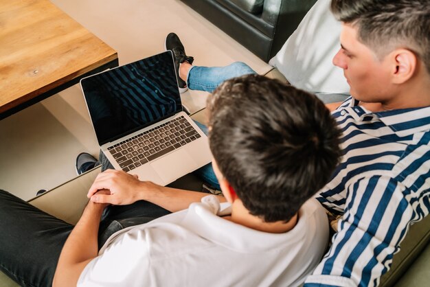 Schwules Paar, das Zeit zusammen hat und einen Laptop benutzt, während es zu Hause auf einer Couch sitzt.