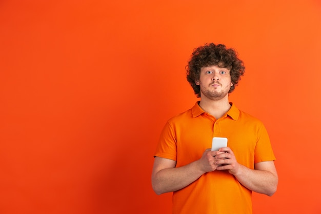 Scroll-Smartphone. Das monochrome Portrait des kaukasischen jungen Mannes an der orangefarbenen Wand. Schönes männliches lockiges Modell im lässigen Stil. Konzept der menschlichen Emotionen, Gesichtsausdruck, Verkauf, Anzeige.