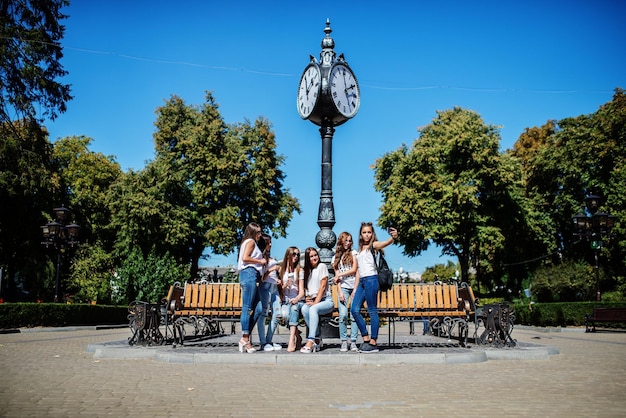 Sechs schöne junge Mädchen sitzen auf einer Bank neben der alten Straßenuhr im Park
