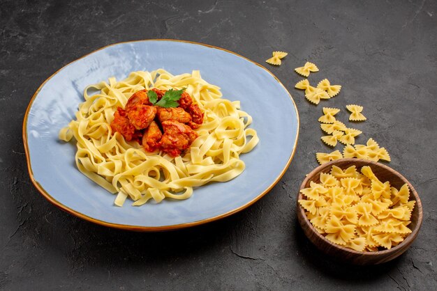 Seitenansicht appetitliche Pasta appetitliche Pasta mit Soße und Fleisch auf dem Teller neben der Schüssel Pasta auf dem dunklen Tisch