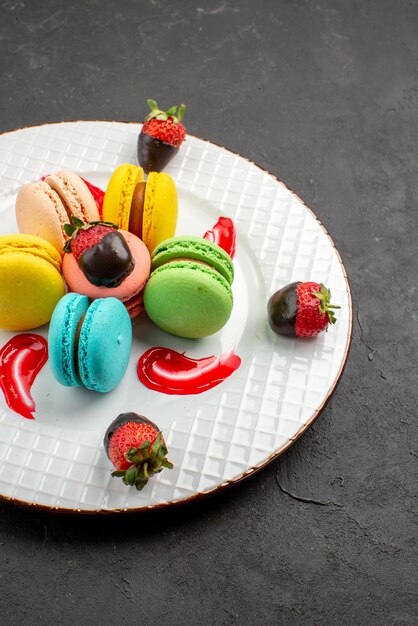 Seitenansicht aus der Ferne bunter Makronenteller mit blau-gelb-rosa und grüner Makronensauce und appetitlichen schokoladenüberzogenen Erdbeeren auf dem dunklen Tisch