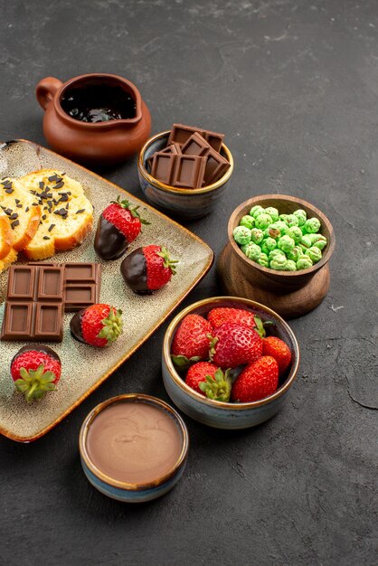 Seitenansicht aus der Ferne Erdbeerkuchen Schokoladenschüsseln mit Schokoladenerdbeeren grüne Bonbons und Schokoladencreme appetitlicher Kuchen und Erdbeeren auf der linken Seite des Tisches