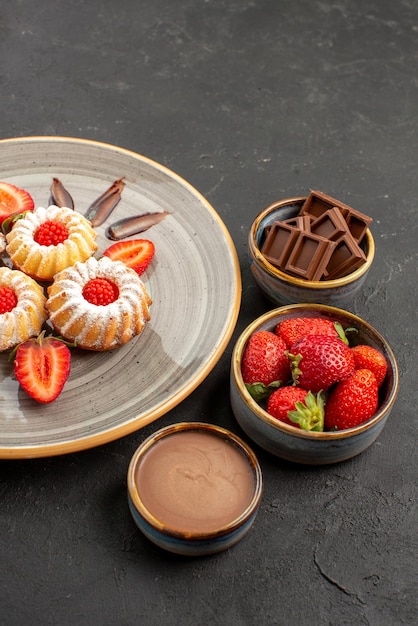 Seitenansicht aus der Ferne Schokoladen- und Keksschalen mit Schokoladenerdbeeren und Schokoladencreme neben dem Teller mit Keksen mit Erdbeeren auf dem Tisch