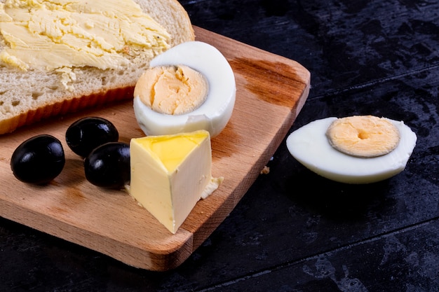 Kostenloses Foto seitenansicht brot- und butterscheiben an bord mit oliven und gekochtem ei auf schwarz