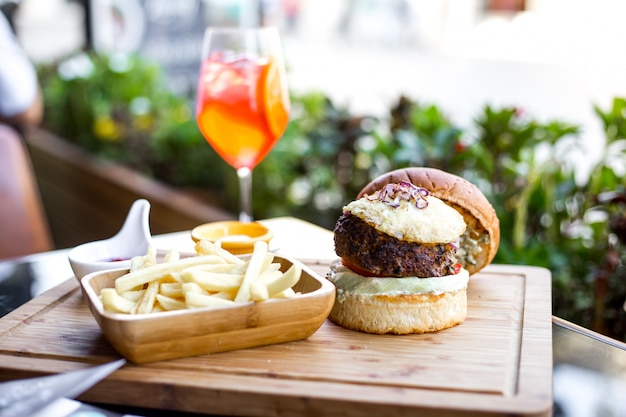 Seitenansicht Burger mit Rindfleischpastetchen gegrilltem Tomaten-Salat der roten Zwiebel in Burgerbrötchen-Pommes Frites und Orangensaftgetränk auf dem Tisch