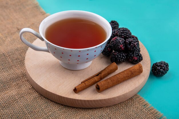 Seitenansicht der Brombeere mit einer Tasse Tee und Zimt auf einem Ständer auf einer türkisfarbenen Oberfläche