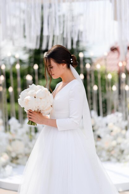 Seitenansicht der charmanten Braut Brünette Frau im Schleier und elegantes Hochzeitskleid mit Blick auf Bouquet von Pfingstrosen, die auf blühenden Altar stehen Trauung im Freien Hübsche Braut