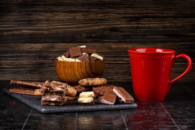 Seitenansicht der dunklen und weißen Schokolade in einer hölzernen Schüssel eine Tasse Tee und Haferkekse verstreut auf dunklem Hintergrund