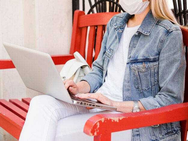 Seitenansicht der Frau im Stuhl, die Gesichtsmaske trägt und am Laptop arbeitet