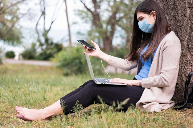 Seitenansicht der Frau mit der medizinischen Maske, die auf Laptop in der Natur arbeitet