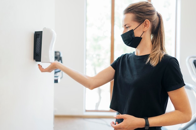 Seitenansicht der Frau mit der medizinischen Maske, die ihre Hände an der Turnhalle desinfiziert